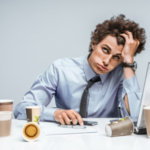 A man looking frustrated in front of a laptop with empty coffee cups scattered across his desk.